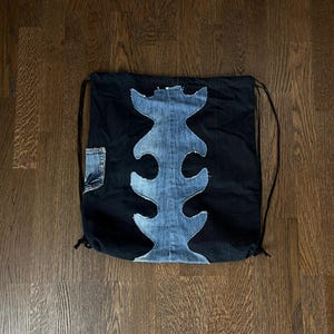 Peut inclure: Sac à cordon noir avec un motif central en denim bleu clair. Le motif en denim a une forme abstraite. Une petite poche en jean est sur le côté. Le sac est sur une surface en bois.