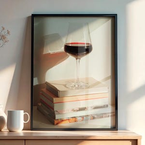 May include: A framed print of a wine glass filled with red wine, resting on a stack of books. A white vase with branches, a small vase, and a white mug sit on a wooden cabinet. The scene is bathed in sunlight.