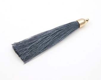 May include: A single, gray tassel with a gold-colored cap. The tassel is made of many thin, silky threads. The cap is a smooth, rounded shape with a small loop at the top. The tassel is set against a white background.