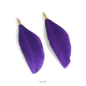 May include: Two purple feather pendants with gold-coloured metal caps. The feathers are a deep, vibrant purple and have a soft, textured appearance. These pendants are likely for jewellery making or craft projects.