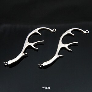 Puede incluir: Dos dijes de metal plateado con forma de astas de ciervo. Cada dije presenta un diseño curvo y ramificado con un pequeño lazo circular en los extremos superior e inferior para su fijación. La palabra WISH está impresa en blanco abajo.