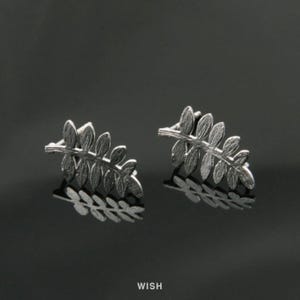 May include: A pair of silver-colored leaf-shaped stud earrings. Each earring features a detailed design of multiple leaves arranged along a stem. The earrings are displayed on a reflective black surface.