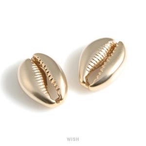 Puede incluir: Dos pendientes dorados con forma de concha de cauri. Los pendientes tienen forma ovalada con una abertura texturizada y estriada. Las conchas tienen una superficie lisa y pulida, sobre un fondo blanco. La palabra "WISH" está en la parte inferior.