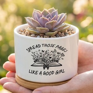 May include: A white ceramic cylindrical planter with a bamboo base, containing a light purple succulent. The planter features a black graphic of an open book with flowers and butterflies, and the text SPREAD THOSE PAGES LIKE A GOOD GIRL.