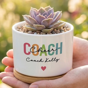 May include: A white ceramic planter with a bamboo base containing a succulent plant. The planter features the colorful text "COACH" in block letters, the cursive word "Cheer," the name "Coach Kelly," and a small red heart printed on the front.