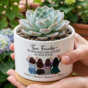 May include: A white ceramic cylindrical planter with a bamboo base containing a light green succulent. The planter features a graphic of four friends with the text True Friends ARE NEVER APART MAYBE IN DISTANCE BUT NEVER IN HEART and names.