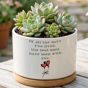 May include: A white ceramic planter with a succulent arrangement. The planter has the text "Of all the days I've lived. the best ones have been with you." and a drawing of two hearts. The planter sits on a wooden base.