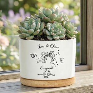 May include: A white ceramic planter with a bamboo base containing a green succulent. The planter features black line art of two hands clinking wine glasses, two rings, and the text: Jesse & Olive, Engaged, 2026, with a small bow graphic.