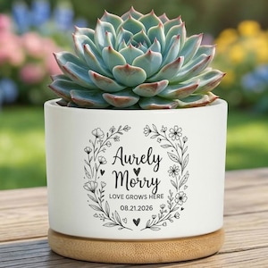 Puede incluir: Una maceta de cerámica blanca para suculentas con base de bambú, que presenta un diseño de corona floral negra alrededor del texto "Aurely Morry", "LOVE GROWS HERE" y la fecha "21.08.2026". Una planta suculenta verde se encuentra en la maceta.