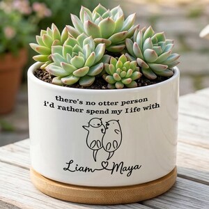May include: A white ceramic cylindrical planter with a bamboo saucer containing a green succulent plant. The planter features black text reading "there's no otter person I'd rather spend my life with" above a line drawing of two otters and the names "Liam & Maya".