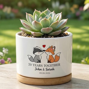Peut inclure: Un pot de fleurs cylindrique en céramique blanche avec une base en bambou contenant une plante succulente verte. L'avant présente un dessin d'un blaireau et d'un renard s'embrassant sous un cœur rouge, avec le texte 20 YEARS TOGETHER, John & Sarah, 08.08.2026.
