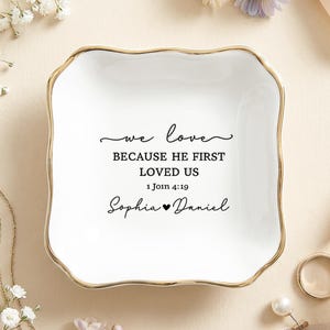 May include: A square white ceramic trinket dish with scalloped edges and a gold-toned rim. The center features black text that reads: we love BECAUSE HE FIRST LOVED US 1 John 4:19 Sophia ♥ Daniel. The dish has a smooth, glossy finish.