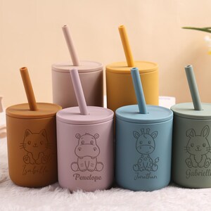 May include: Six silicone cups in various pastel shades, each with a lid and straw. The cups are decorated with animal illustrations and names. Colours include beige, pink, yellow, blue, and green. Designed for children.
