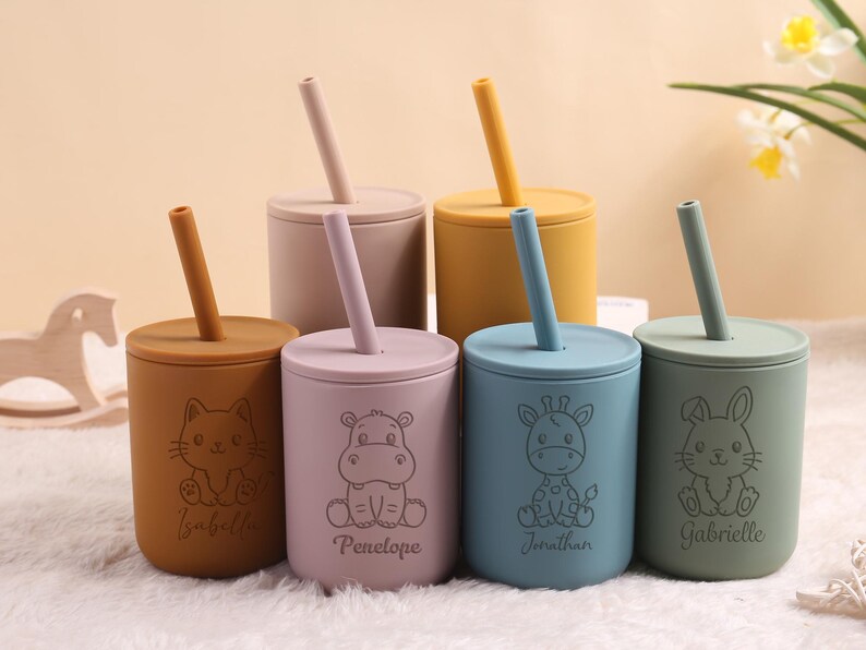 May include: Six silicone cups in various pastel shades, each with a lid and straw. The cups are decorated with animal illustrations and names. Colours include beige, pink, yellow, blue, and green. Designed for children.