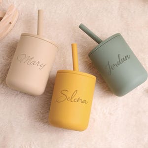 May include: Three personalised baby cups with straws in beige, yellow, and green. Each cup has a name engraved on the front: Mary, Selena, and Jordan. The cups are made of a soft, matte material, ideal for little hands.