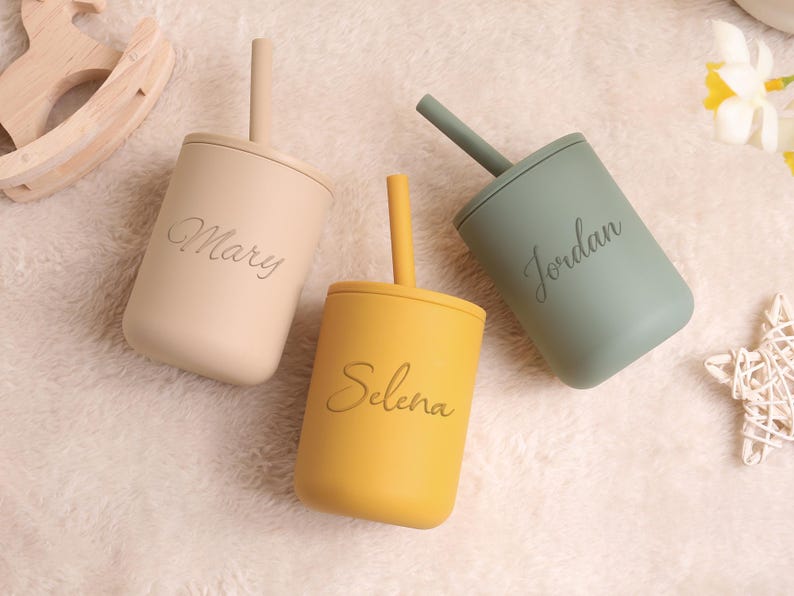 May include: Three personalised baby cups with straws in beige, yellow, and green. Each cup has a name engraved on the front: Mary, Selena, and Jordan. The cups are made of a soft, matte material, ideal for little hands.