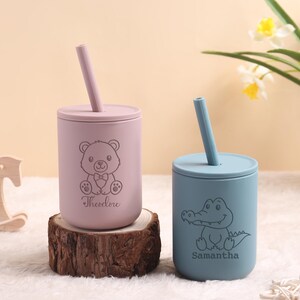 May include: Two personalised baby cups with straws. One is light purple with a teddy bear design and the name "Theodore". The other is light blue with an alligator design and the name "Samantha". Both cups are on a wooden base.