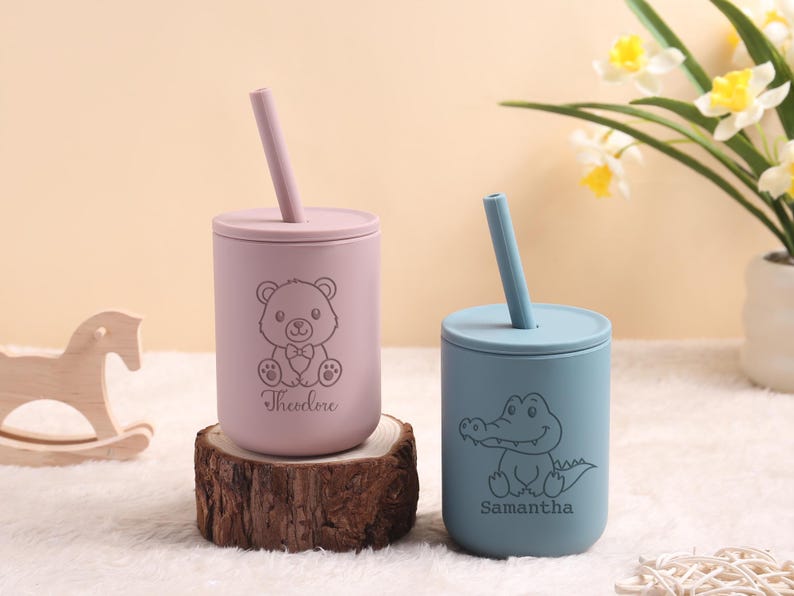 May include: Two personalised baby cups with straws. One is light purple with a teddy bear design and the name "Theodore". The other is light blue with an alligator design and the name "Samantha". Both cups are on a wooden base.