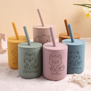 May include: A set of colourful silicone cups with lids and straws. Each cup has a different animal illustration, such as a tiger, fox, cat, and elephant. The cups are personalised with names like "Alexander" and "Jonathan".
