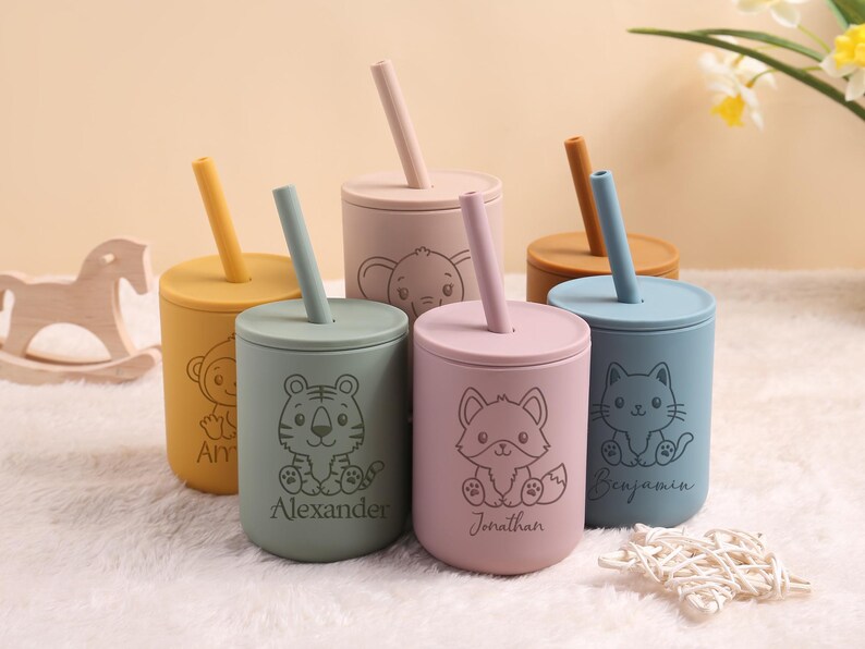 May include: A set of colourful silicone cups with lids and straws. Each cup has a different animal illustration, such as a tiger, fox, cat, and elephant. The cups are personalised with names like "Alexander" and "Jonathan".