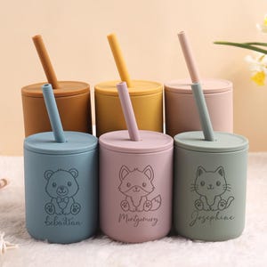 May include: Six colourful silicone cups with lids and straws. Each cup features a different animal design: a bear, fox, and cat, with a name below each. The cups are in shades of blue, pink, and green, with matching straws.