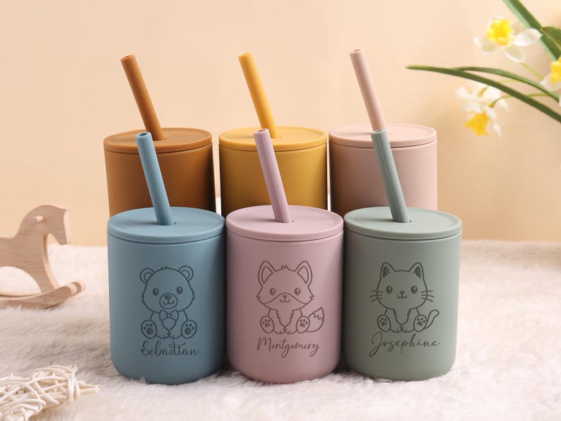 May include: Six colourful silicone cups with lids and straws. Each cup features a different animal design: a bear, fox, and cat, with a name below each. The cups are in shades of blue, pink, and green, with matching straws.
