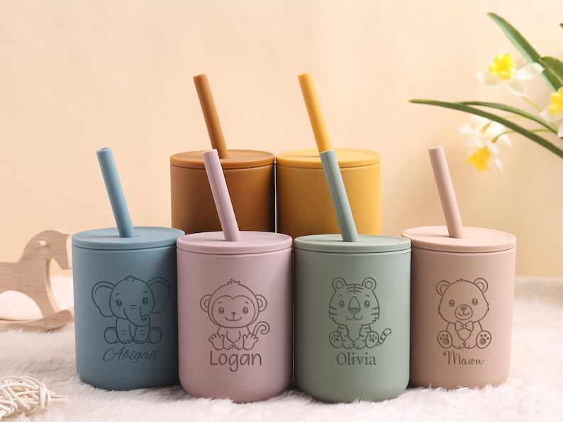 May include: A collection of six colorful silicone cups with lids and straws. Each cup features a different animal illustration and a name. The cups are in shades of blue, pink, green, and tan, with matching straws.