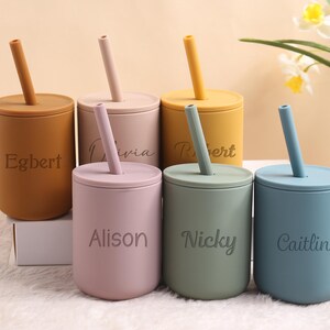 May include: Six pastel-coloured silicone cups with lids and straws. The colours are mustard, tan, pink, yellow, sage green, and blue. Each cup has a name engraved on it: Egbert, Olivia, Robert, Alison, Nicky, and Caitlin.