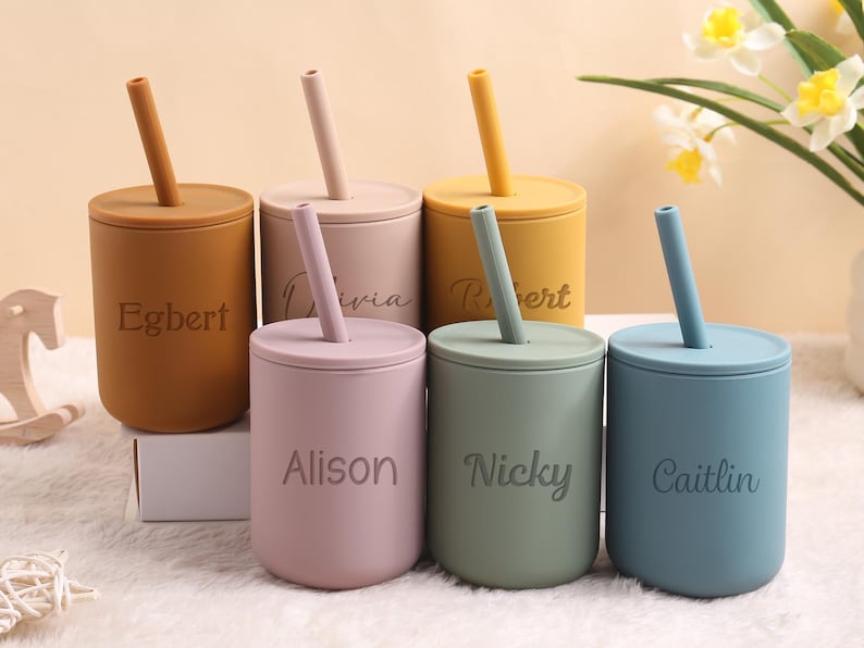 May include: Six pastel-coloured silicone cups with lids and straws. The colours are mustard, tan, pink, yellow, sage green, and blue. Each cup has a name engraved on it: Egbert, Olivia, Robert, Alison, Nicky, and Caitlin.