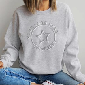 Puede incluir: Una sudadera gris claro de cuello redondo con un diseño circular bordado en el pecho. El diseño incluye una estrella central de cinco puntas rodeada por un borde circular con el texto YOUR LOGO HERE y un motivo de hojas de laurel abajo.