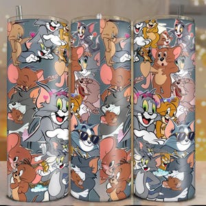 May include: A set of three stainless steel tumblers featuring a repeating pattern of cartoon characters Tom and Jerry. The design includes various poses of the cat and mouse duo in shades of gray, brown, and pink, set against a gray background.