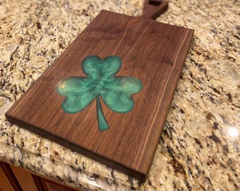 Può includere: Tagliere rettangolare in legno con manico. La tavola presenta al centro un disegno a trifoglio verde. Il legno è di un ricco colore marrone scuro e il trifoglio ha un aspetto lucido e marmorizzato.