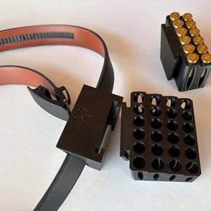 Può includere: Una cintura nera con interno marrone e fibbia nera. Accanto alla cintura ci sono due supporti neri, uno pieno di proiettili dorati e l'altro con fori vuoti.