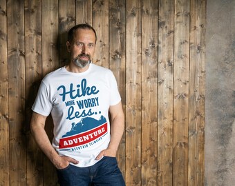 May include: White t-shirt with the text "Hike more worry less.. ADVENTURE MOUNTAIN CLIMBING" in blue and red. The shirt features a mountain graphic. The person is wearing blue jeans and is leaning against a wooden wall.