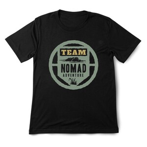 May include: Black t-shirt with a circular graphic. The graphic features the words "TEAM NOMAD ADVENTURE" in a stylized design with a nature theme. The text is in a combination of tan and green colors.