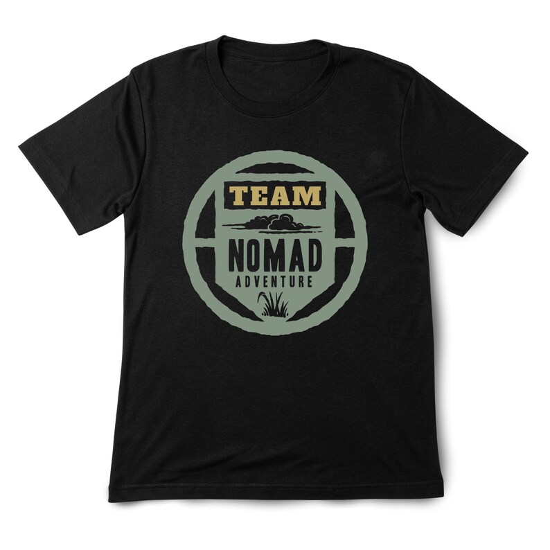 May include: Black t-shirt with a circular graphic. The graphic features the words "TEAM NOMAD ADVENTURE" in a stylized design with a nature theme. The text is in a combination of tan and green colors.