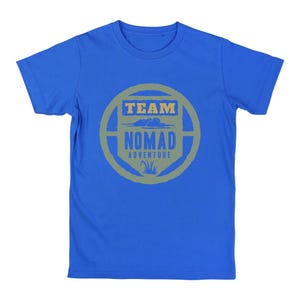 May include: A blue t-shirt with a circular graphic in shades of gray and green. The graphic reads "TEAM NOMAD ADVENTURE" with a cloud and grass design. The shirt is made of a soft material.