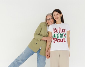 May include: A white t-shirt with the text "Better NOT Pout" in red and green, with a Christmas tree and holly graphic. The shirt is worn by a person standing next to another person wearing a green blazer and blue jeans.