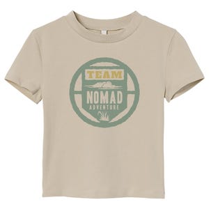 May include: A tan crew neck t-shirt featuring a circular graphic with the words "TEAM NOMAD ADVENTURE" in green and gold. The design includes cloud and grass illustrations. The shirt is made of a soft material.