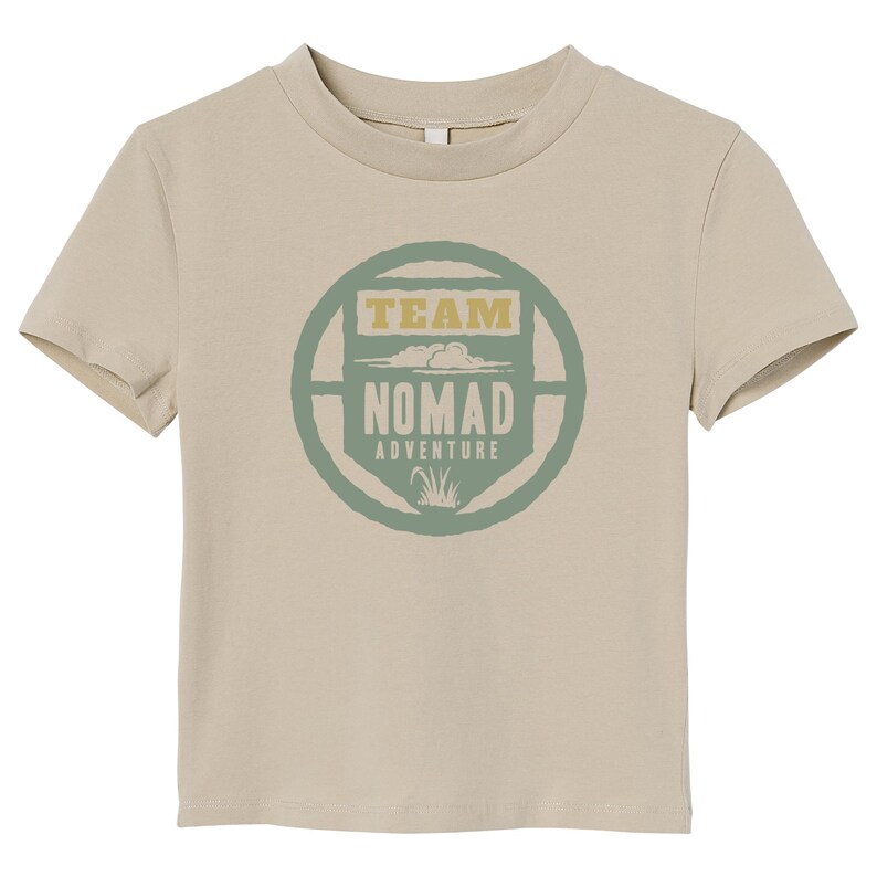 May include: A tan crew neck t-shirt featuring a circular graphic with the words "TEAM NOMAD ADVENTURE" in green and gold. The design includes cloud and grass illustrations. The shirt is made of a soft material.