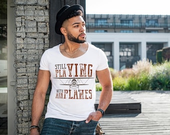 May include: White t-shirt with the text "STILL PLAYING WITH AIRPLANES" in brown lettering and a graphic of a biplane. The person is wearing a black hat and blue jeans.