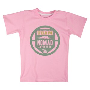 May include: A light pink t-shirt with a crew neck and short sleeves. The shirt features a circular design in green and tan with the words "TEAM NOMAD ADVENTURE" in a stylized font. The design includes cloud and grass graphics.