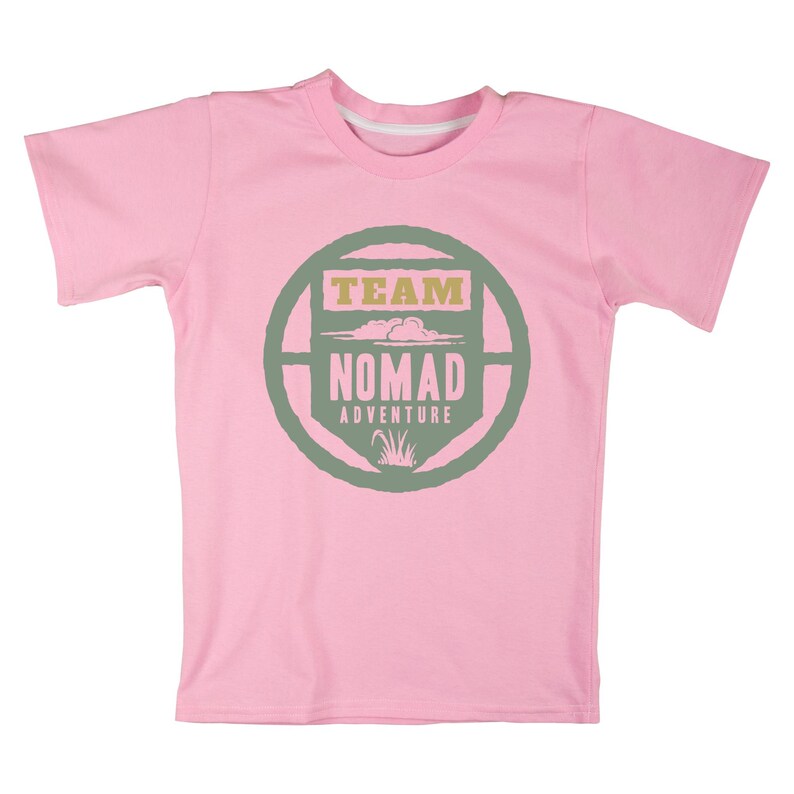 May include: A light pink t-shirt with a crew neck and short sleeves. The shirt features a circular design in green and tan with the words "TEAM NOMAD ADVENTURE" in a stylized font. The design includes cloud and grass graphics.