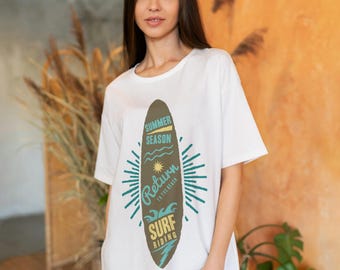 May include: White t-shirt featuring a surfboard design with the text "Summer Season, Return to the Beach, Surf Riding". The graphic is in shades of green, blue, and brown. The shirt is a casual, relaxed fit.