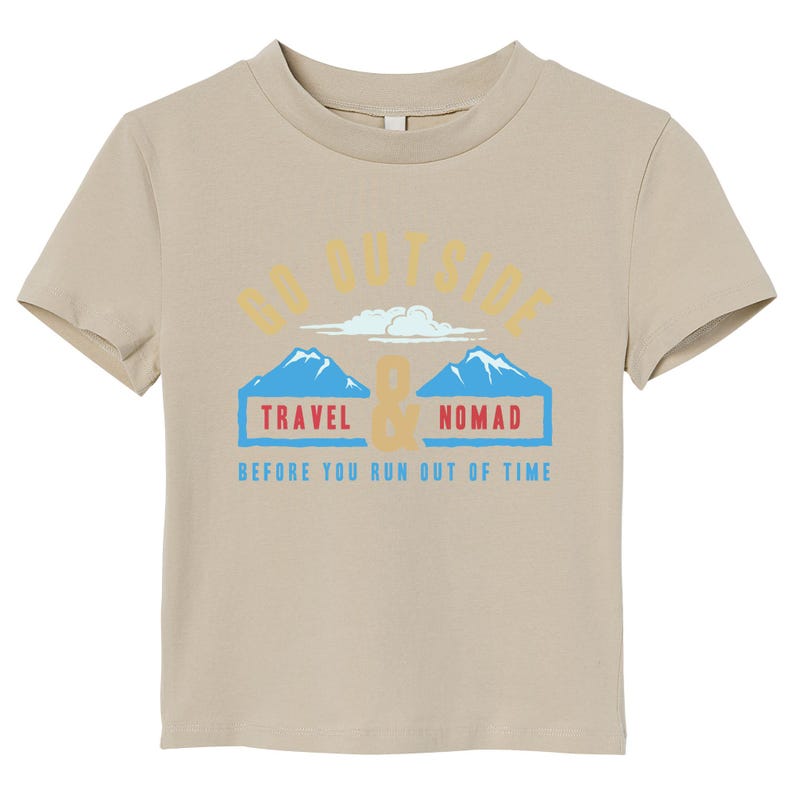 May include: A tan-colored t-shirt with a crew neck. The shirt features the text "GO OUTSIDE" above a graphic of two blue mountains and the words "TRAVEL & NOMAD" in blue boxes. Below is the text "BEFORE YOU RUN OUT OF TIME".