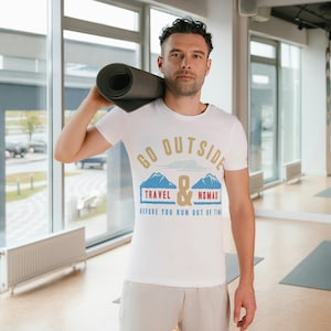 May include: A white t-shirt with the text "GO OUTSIDE TRAVEL & NOMAD before you run out of time" in gold, blue, and red. The person is holding a rolled-up yoga mat. The setting appears to be a gym or studio.