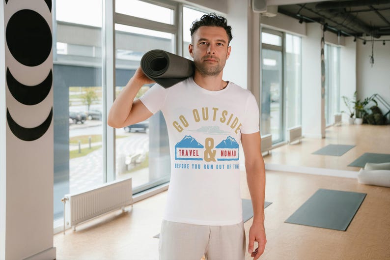 May include: A white t-shirt with the text "GO OUTSIDE TRAVEL & NOMAD before you run out of time" in gold, blue, and red. The person is holding a rolled-up yoga mat. The setting appears to be a gym or studio.