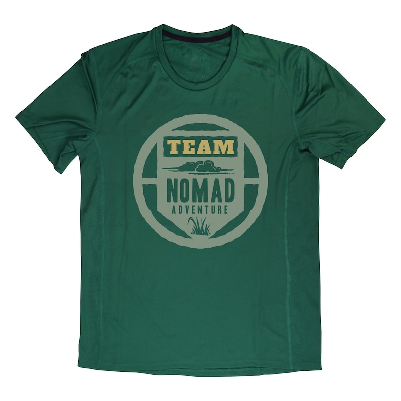 May include: A dark green athletic t-shirt with a crew neck and short sleeves. The shirt features a circular graphic with the words "TEAM NOMAD ADVENTURE" in a light tan color. The graphic includes a cloud and grass design.