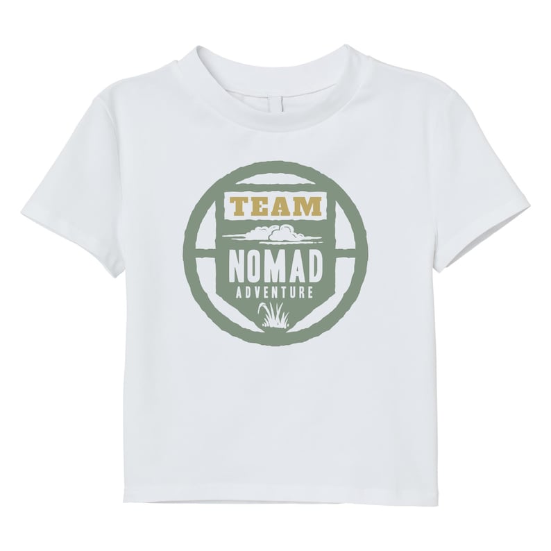 May include: White t-shirt with a crew neck and short sleeves. The front features a circular design with the words "TEAM NOMAD ADVENTURE" in a green and tan color scheme. The design includes cloud and grass graphics.