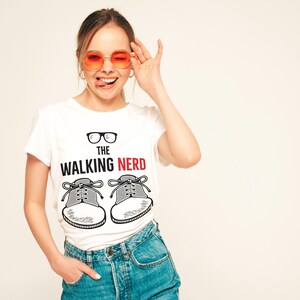 Pode incluir: Camiseta branca com o texto "THE WALKING NERD" e um gráfico de óculos e tênis. A pessoa está usando óculos de sol laranja e jeans azuis.